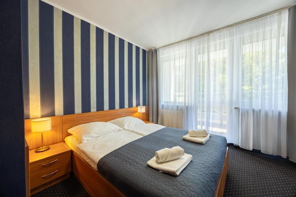 Ein Hotelzimmer mit einem großen Bett, einem Nachttisch mit Lampe und einem Fenster mit transparenten Vorhängen.