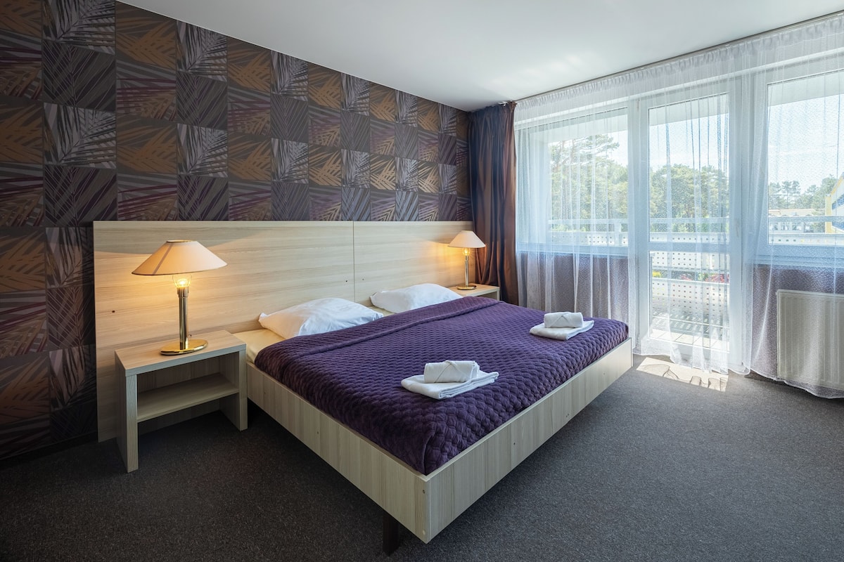 A hotel room with a large bed, two bedside tables with lamps, a patterned wall, and a view through sheer curtains.