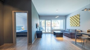 A modern living room with a sofa, dining table, and chairs. There is a bed in the adjacent room, and large windows provide natural light.