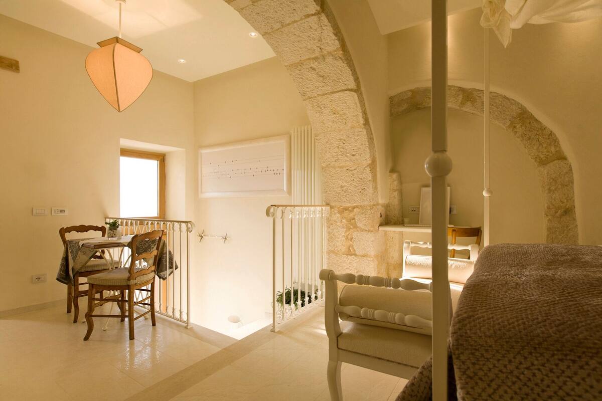 A room with a stone wall, a bed, a chair, a table with chairs, and a hanging light fixture.