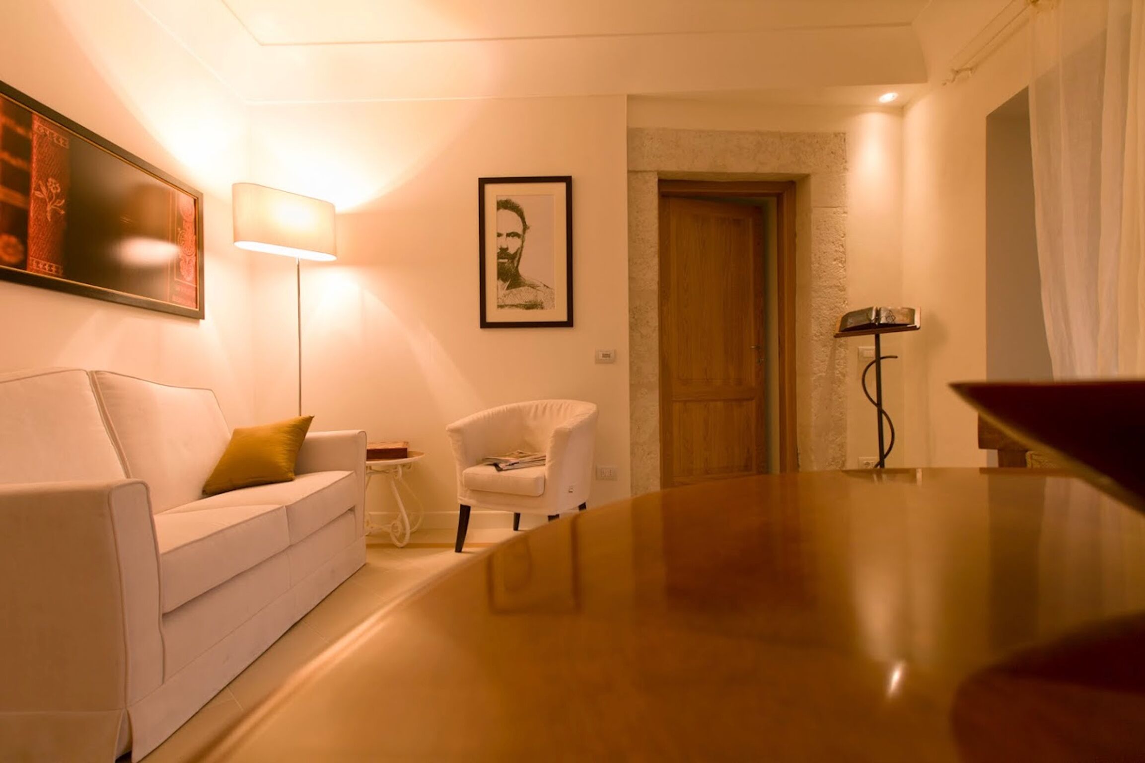 A modern hotel room with a white sofa, a small white armchair, a wooden coffee table, and a framed portrait on the wall.
