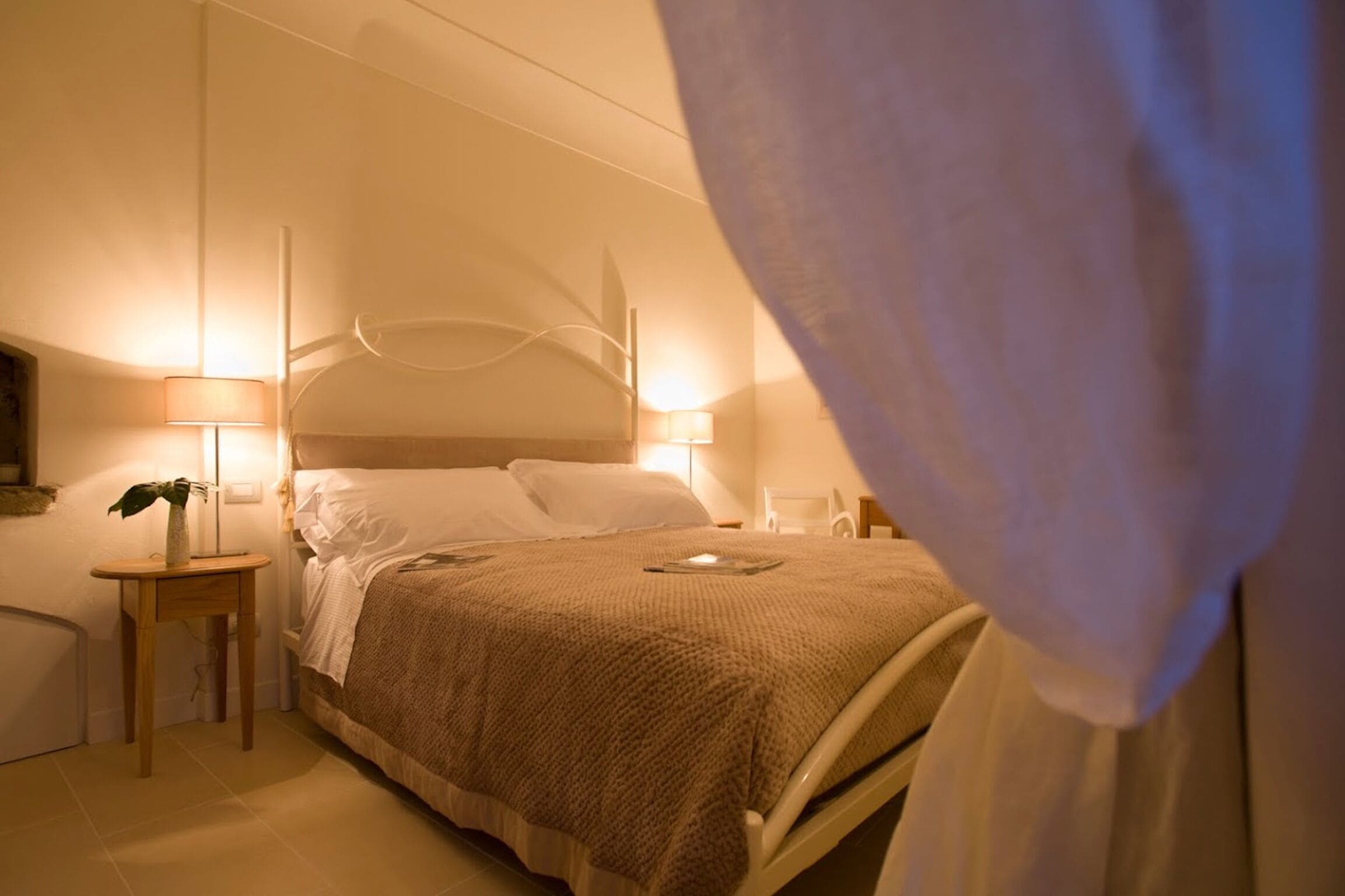 Un dormitorio con cama, lámparas de noche, una mesita de noche con una planta y una cortina entreabierta.