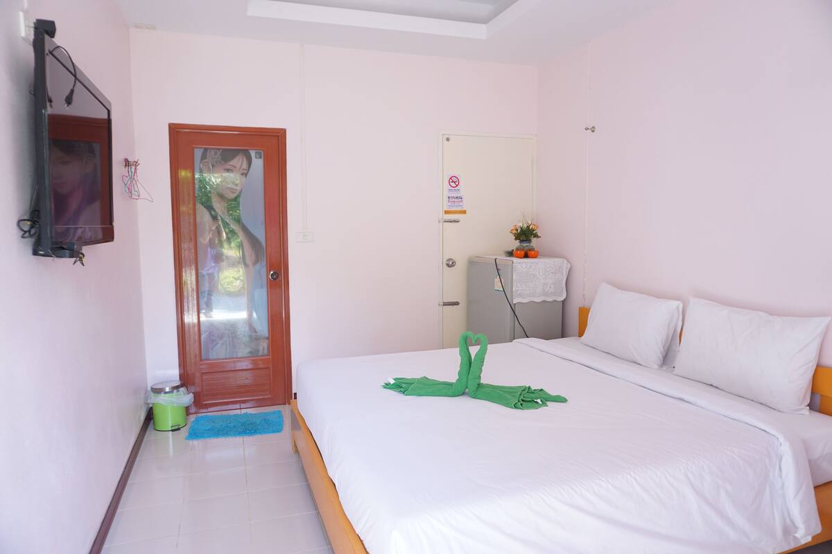 A hotel room with a bed, a door, a mirror, a small table with a plant, and a trash bin.