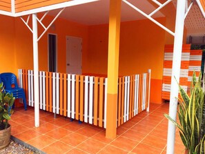 Un espacio exterior techado con paredes naranja, una cerca blanca y naranja, una silla azul y una planta en maceta.