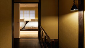 Corredor estreito que dá para um quarto com uma cama, mesa de cabeceira e janela com persianas.