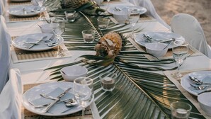 A beachside dining table set with white tablecloths, plates, cutlery, and glasses, surrounded by palm leaves and a pineapple centerpiece.