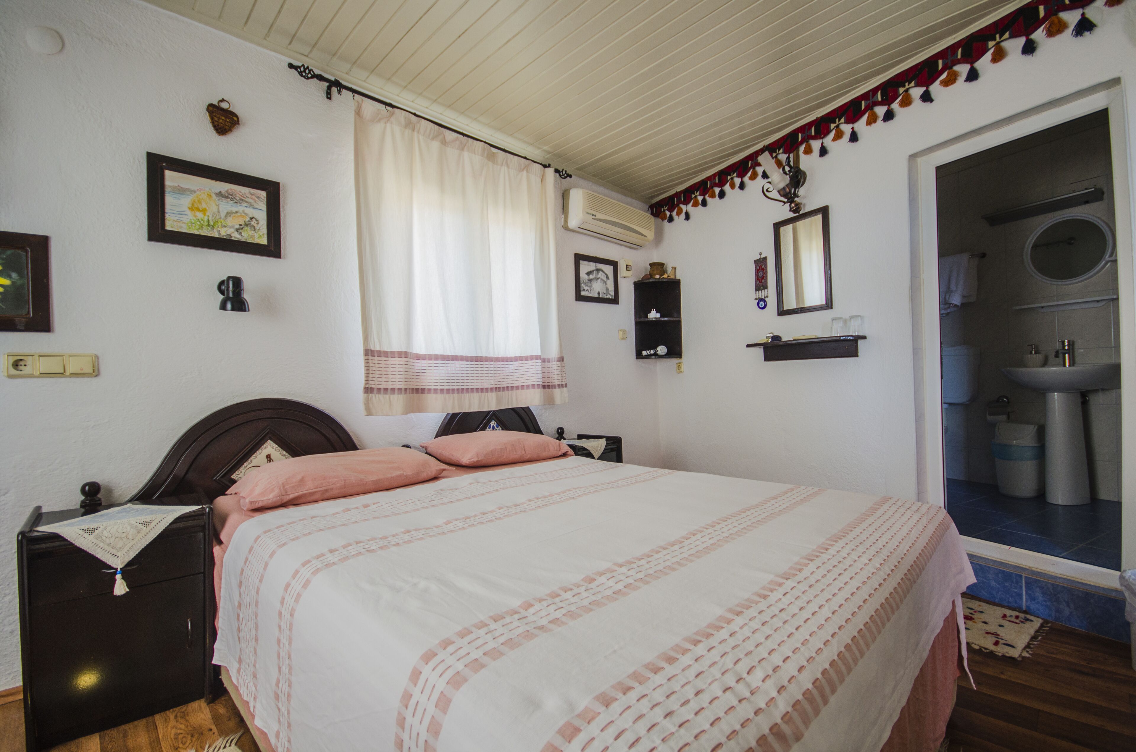Ein Schlafzimmer mit Bett, Nachttisch, Kommode, einem Fenster mit Vorhängen, einem an der Wand befestigten Regal mit Gegenständen und einem durchgegangenen Tür sichtbar ersichtlichen Badezimmer.