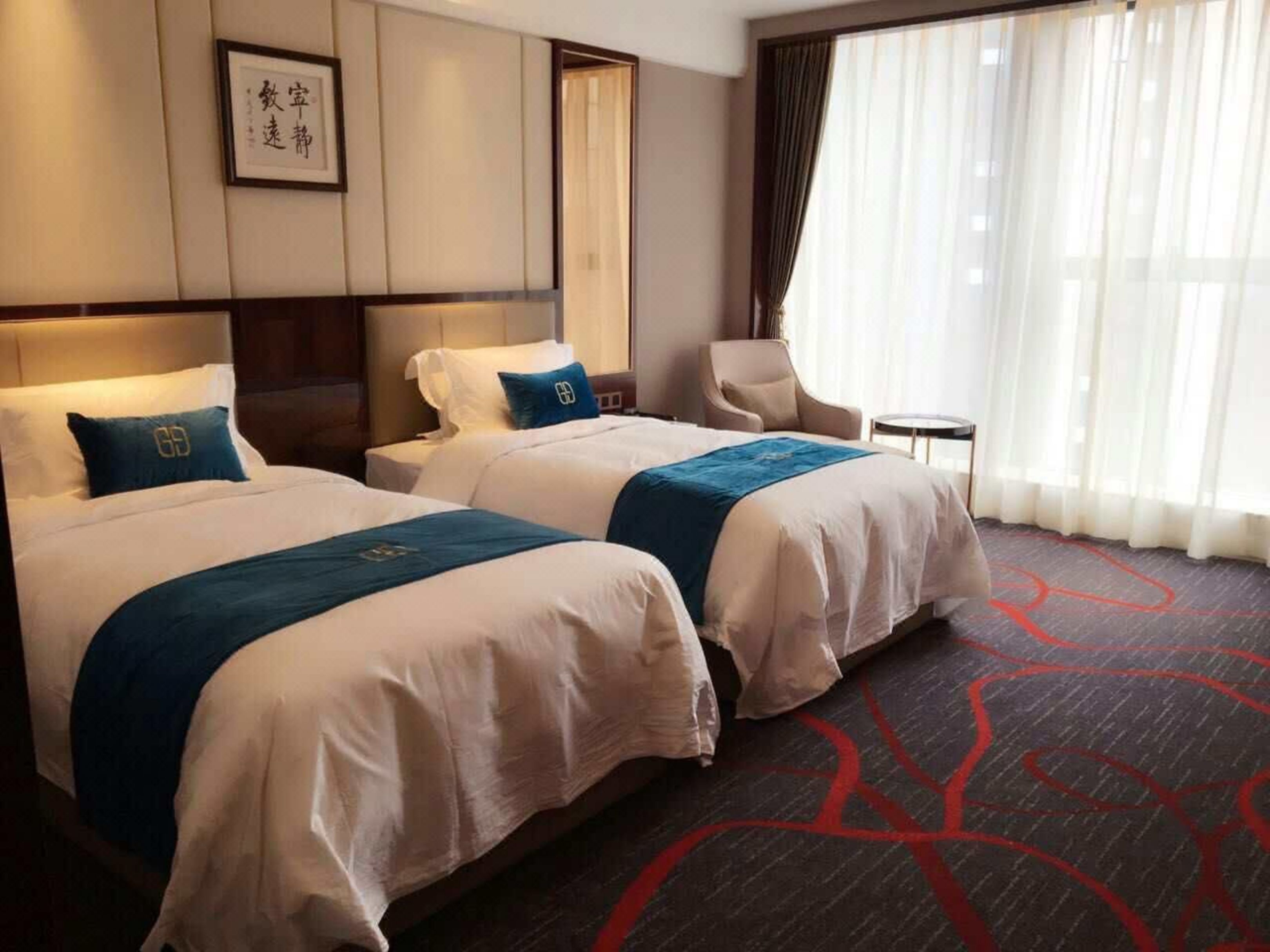 Ein Hotelzimmer mit zwei Betten, einem Sessel, einem kleinen Tisch und einem großen Fenster mit Vorhängen.