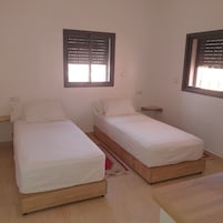 Two single beds with wooden frames and white bedding, a small red rug, and a window with blinds.
