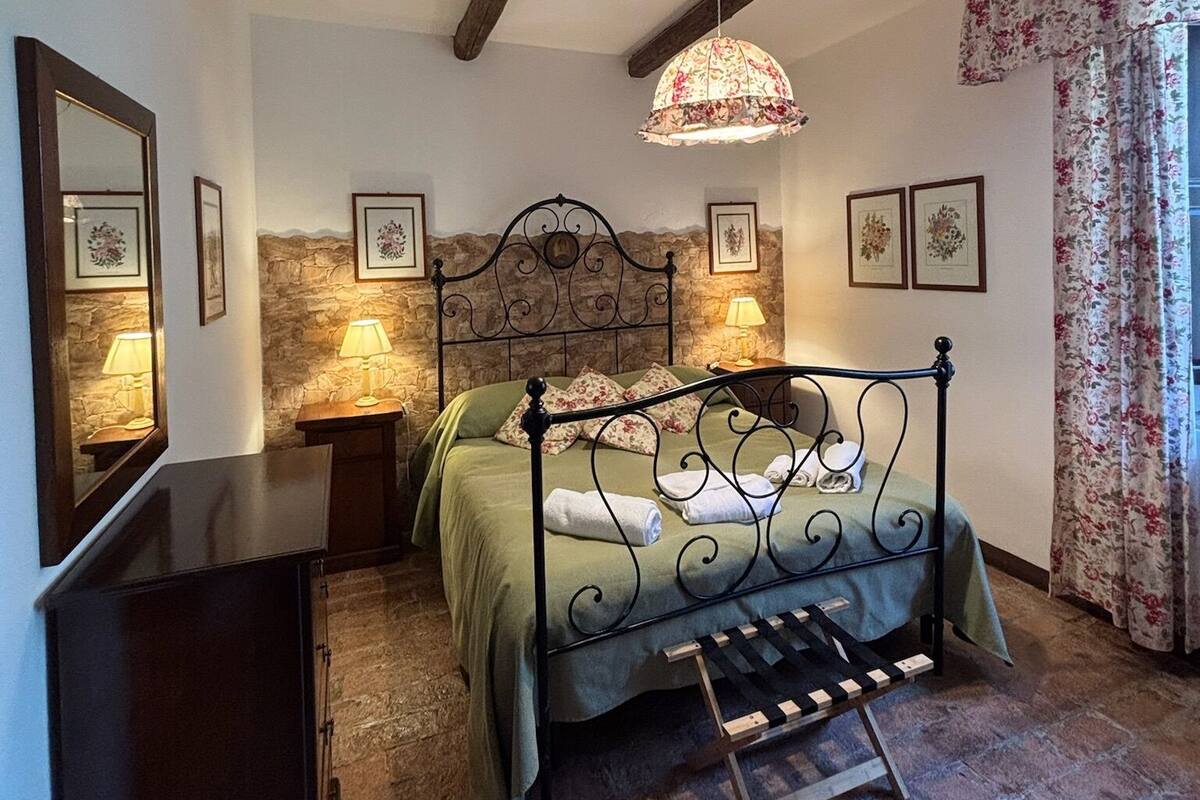 A bedroom with a stone wall, a bed with a metal frame, a wooden bench, and a hanging light fixture.
