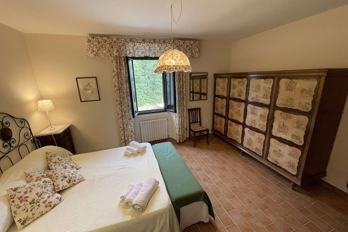 A bedroom with a bed, a radiator, a window with curtains, a chair, a wardrobe, and a hanging lamp.