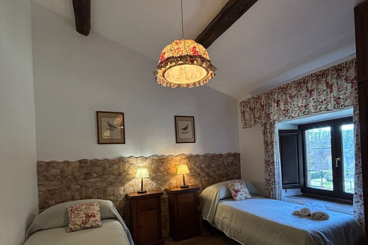 A cozy room with two beds, a stone wall, wooden beams, a hanging lamp, and a window with curtains.