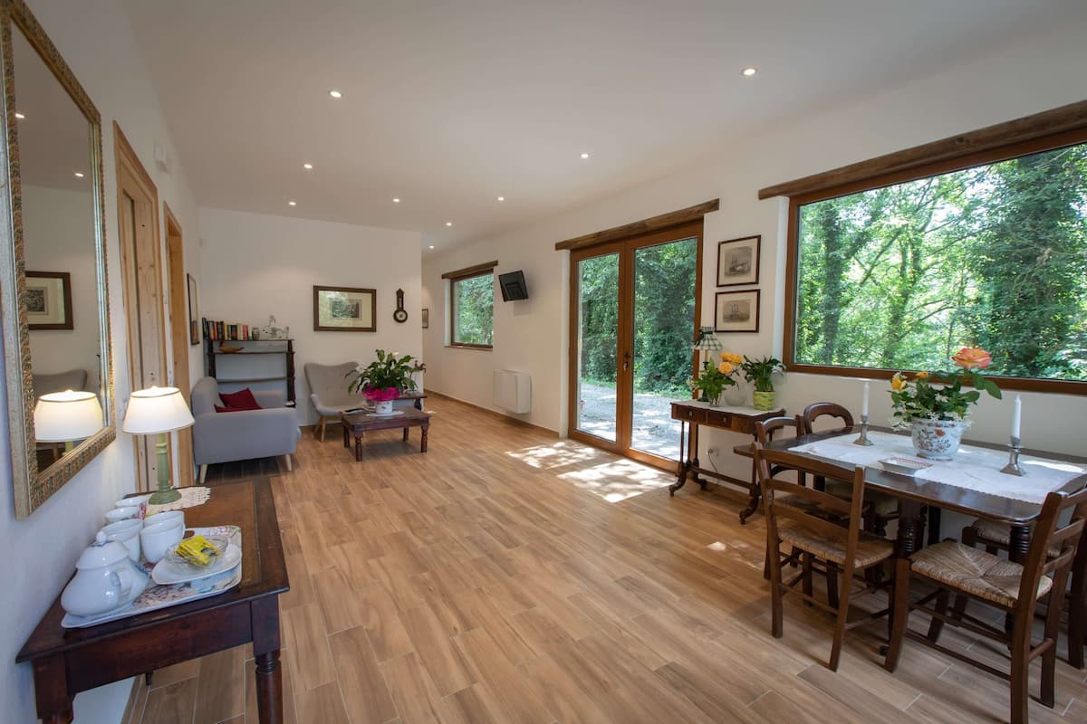 A spacious living area with a wooden floor, a dining table with chairs, a sofa, and a coffee table. There are framed pictures on the walls and a large window with a view of trees.