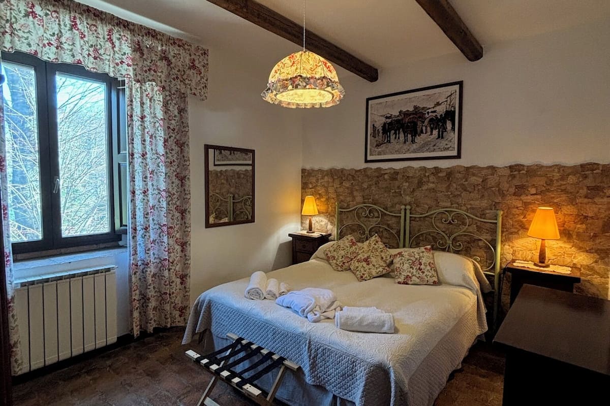 A cozy bedroom with a stone wall, a bed with floral bedding, a hanging lamp, and a radiator.