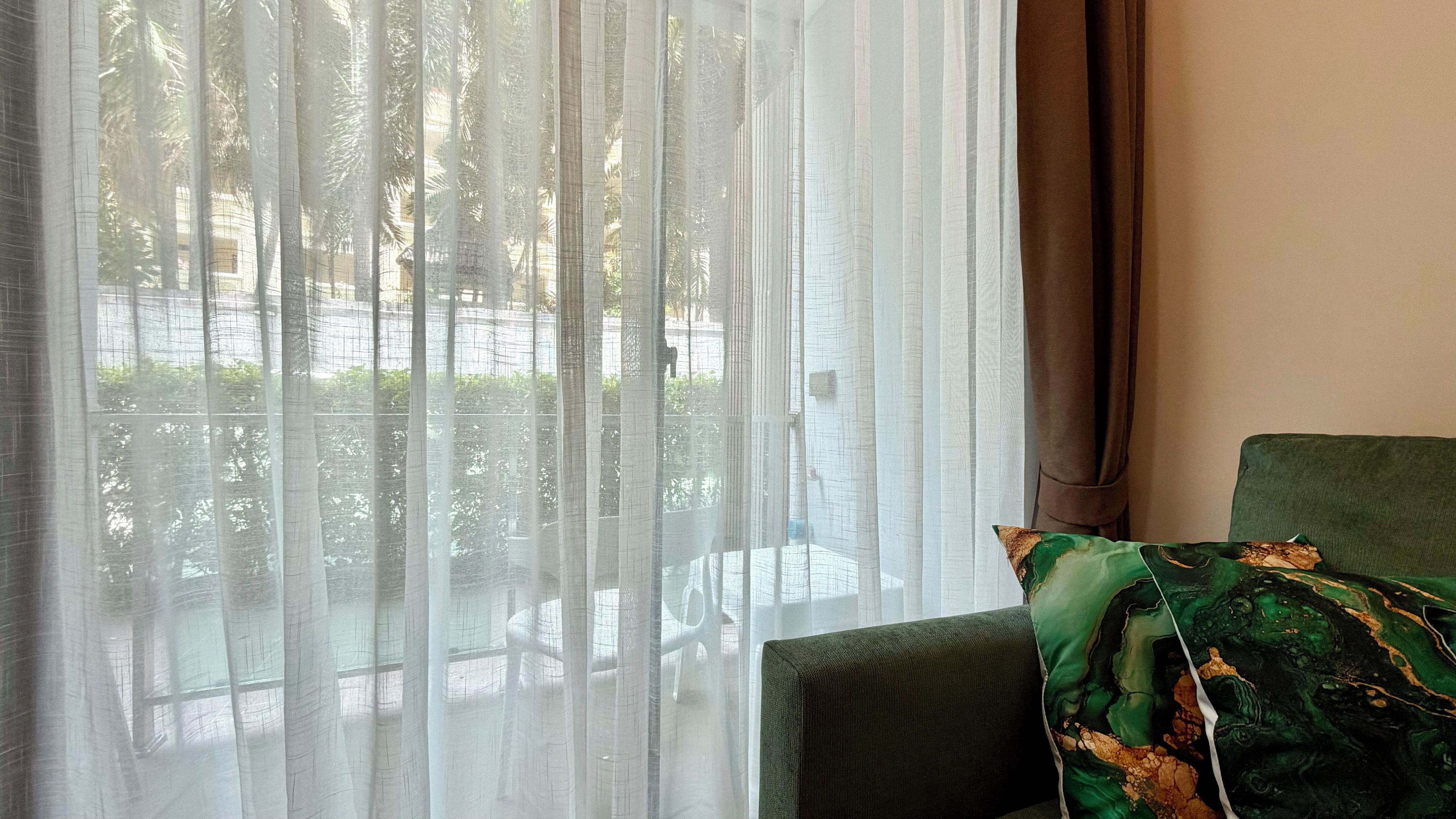 A room with a view through sheer curtains, a green upholstered chair, and a patterned pillow.