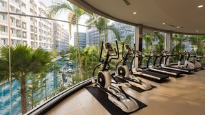 Un gimnasio con bicicletas estáticas y cintas de correr, ventanas amplias con vista a una piscina y palmeras, y un edificio moderno al fondo.