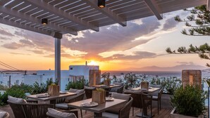 An outdoor dining area with tables and chairs, overlooking a scenic view of the sea and mountains during sunset. - Oniro Suites (Mykonos)