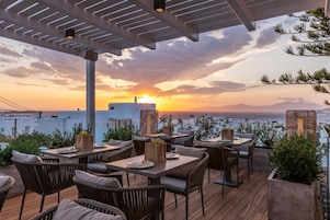 Un área de comedor al aire libre con mesas y sillas, con vistas a un paisaje pintoresco del mar y las montañas durante el atardecer.