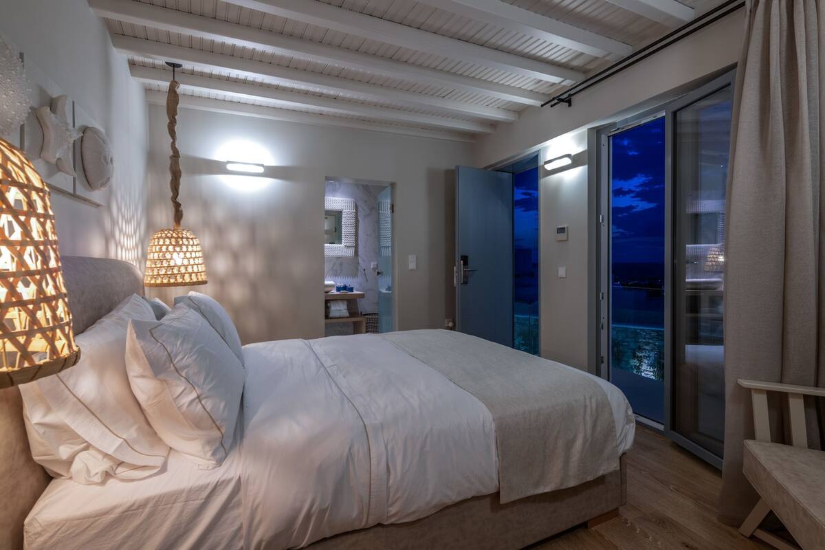 Quarto com cama, luminária pendente, porta para uma varanda e vista para o mar.