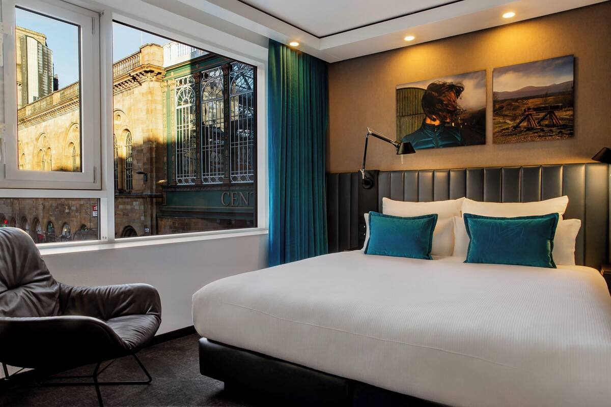 A hotel room with a large bed, a chair, a window with a view of a building, and two paintings on the wall.