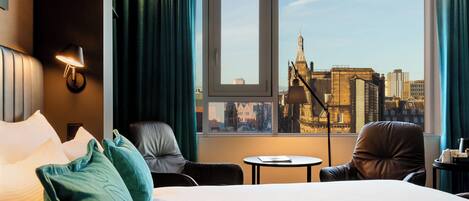 A hotel room with a large bed, two chairs, a small table, and a window with a city view.
