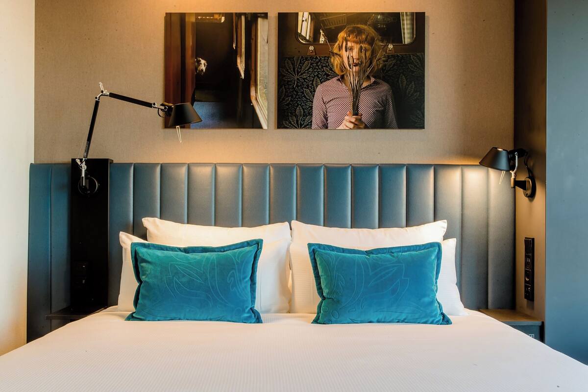 A hotel room with a large bed, two pillows, a headboard, a nightstand, a lamp, and a painting on the wall.