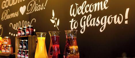 Una pared con los carteles "Welcome to Glasgow!" y "Good Morning", junto con una exhibición de bebidas coloridas y frascos de mermelada.