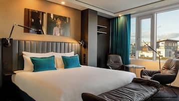 A hotel room with a large bed, two chairs, a small table, and a view of the city.