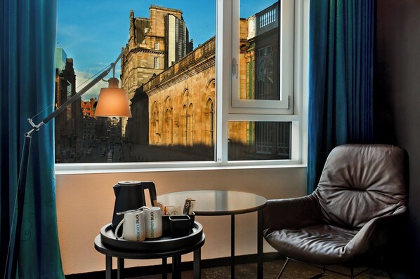 A room with a view of historic buildings, a leather chair, a small table with a lamp, and a tray with a coffee pot and a mug. - Motel One Glasgow (Glasgow)