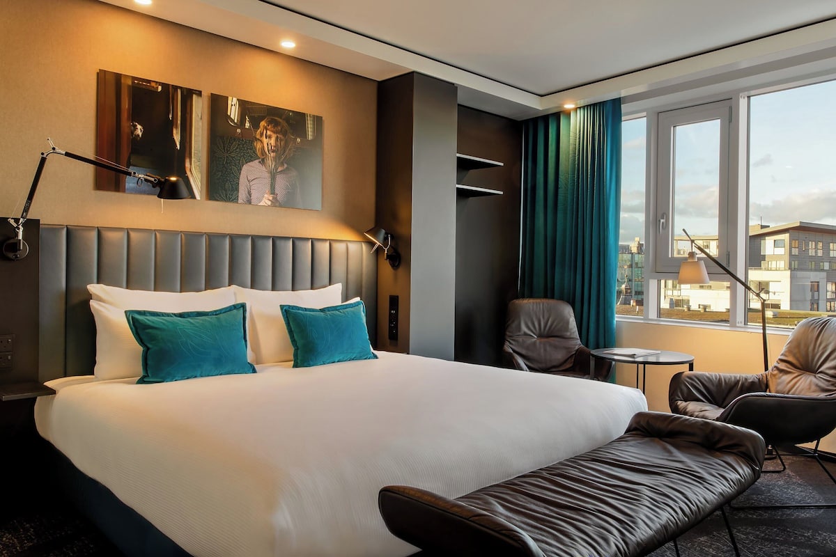 A hotel room with a large bed, a desk, a chair, and a view of the city.