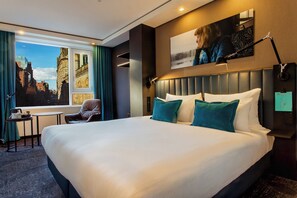 Habitación de hotel con una cama grande, vistas de la ciudad a través de una ventana, una mesita con una silla y una lámpara de pared.