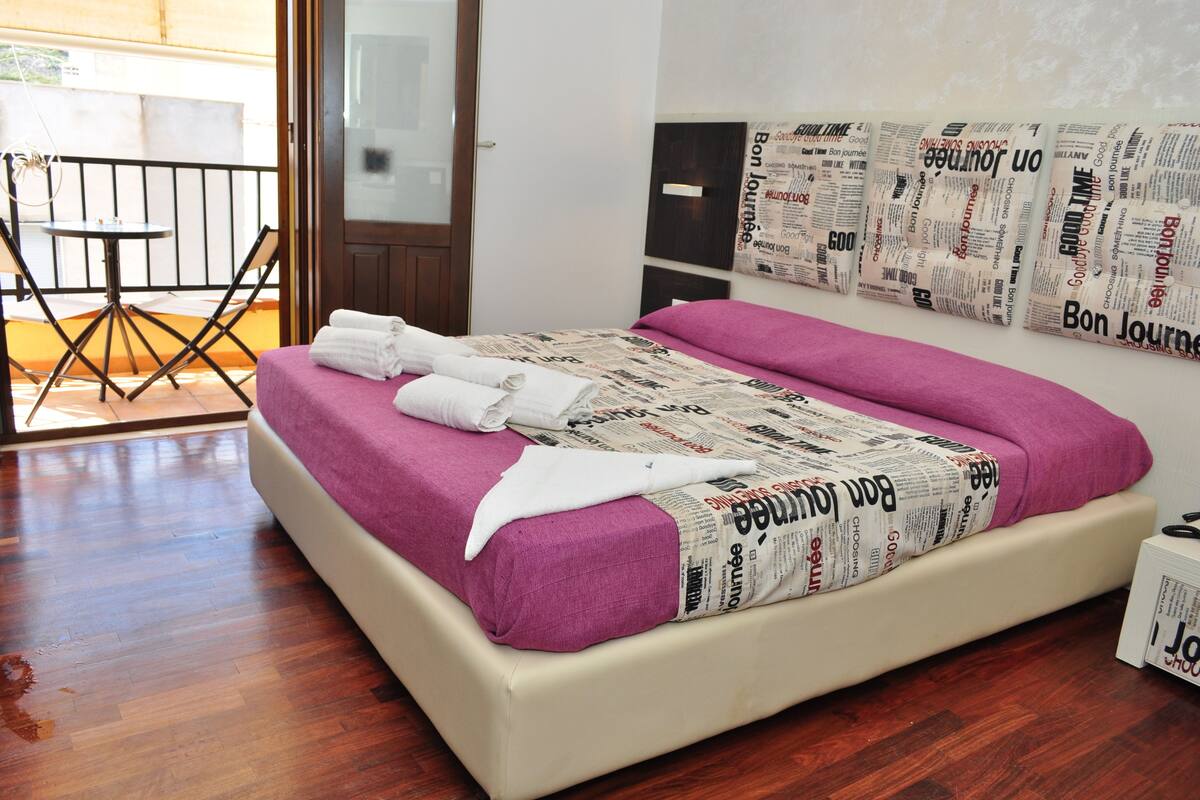 Una cama con un cubrecolcha de periódico, una mesita con sillas y una puerta que da a un espacio exterior.