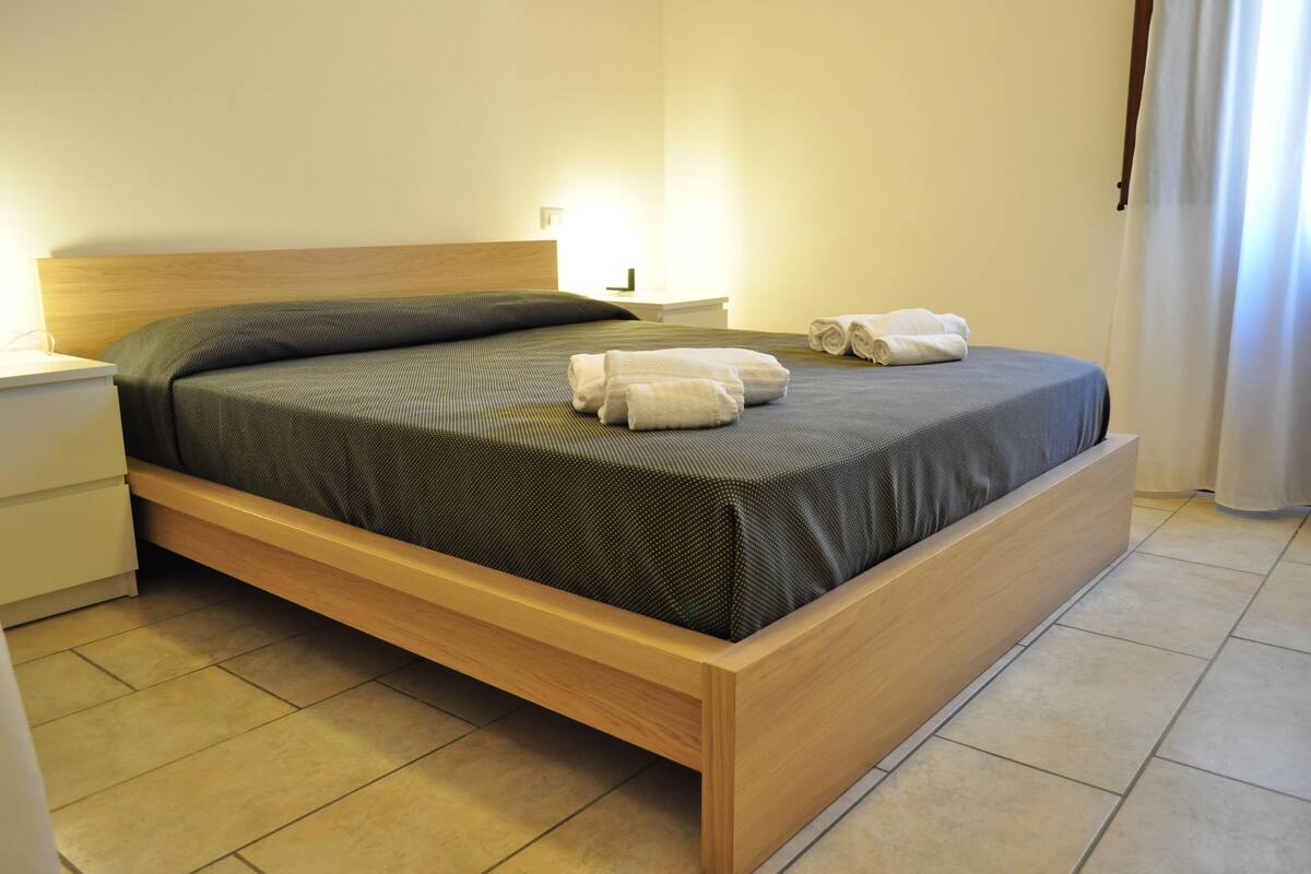 Una cama bien tendida con estructura de madera y colcha verde, junto a dos toallas enrolladas a un lado.