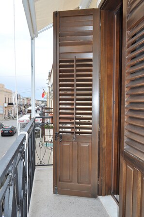 A wooden door with horizontal slats, a balcony with a metal railing, and a street view with buildings and a flag. - Zefiro Rooms & Breakfast (San Vito Lo Capo)
