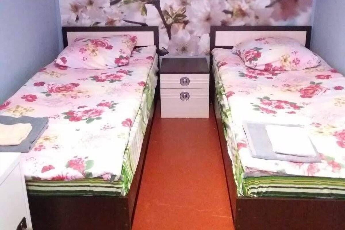 Une chambre avec deux lits, chacun agrémenté de draps à motifs floraux, une table de chevet avec des tiroirs et une fresque murale représentant des fleurs de cerisier.
