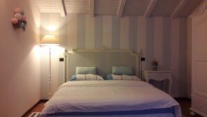 A bed with a white and blue bedspread, a white headboard, and two pillows. There is a bedside table with a lamp and a small plant. The room has striped wallpaper and a wooden ceiling. - B&B Lake Como Cottage (Lecco)