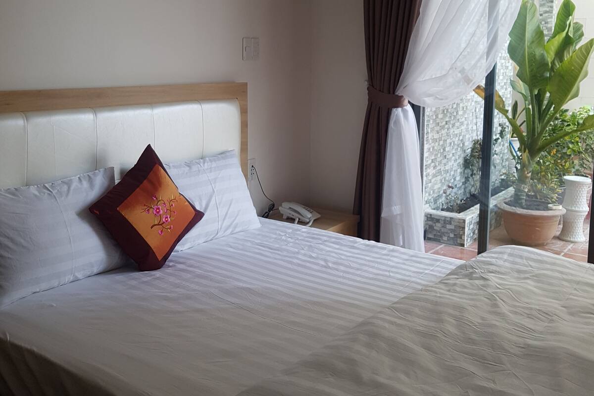 A neatly made bed with white linens and a single decorative pillow, a bedside phone, and a window with sheer curtains.