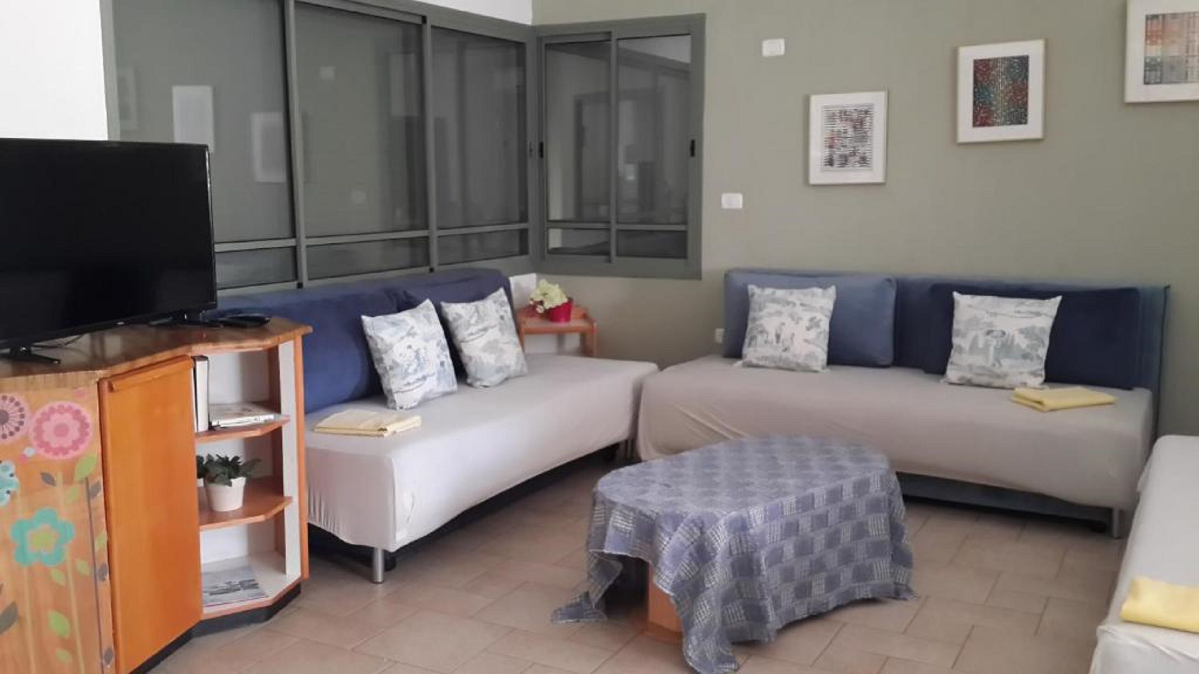 Uma sala de estar com televisão sobre um móvel de madeira, dois sofás com almofadas estampadas, uma mesa pequena com um vaso vermelho e um banquinho estampado.