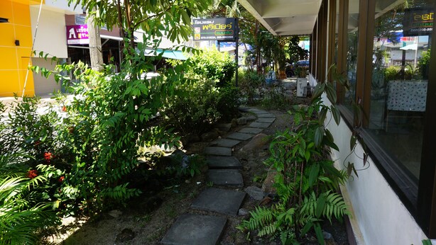 A stone pathway leading through a lush garden area with various plants and a building with signage in a foreign language. - Premier Hotel Nakhonsithammarat (Nakhon Si Thammarat)