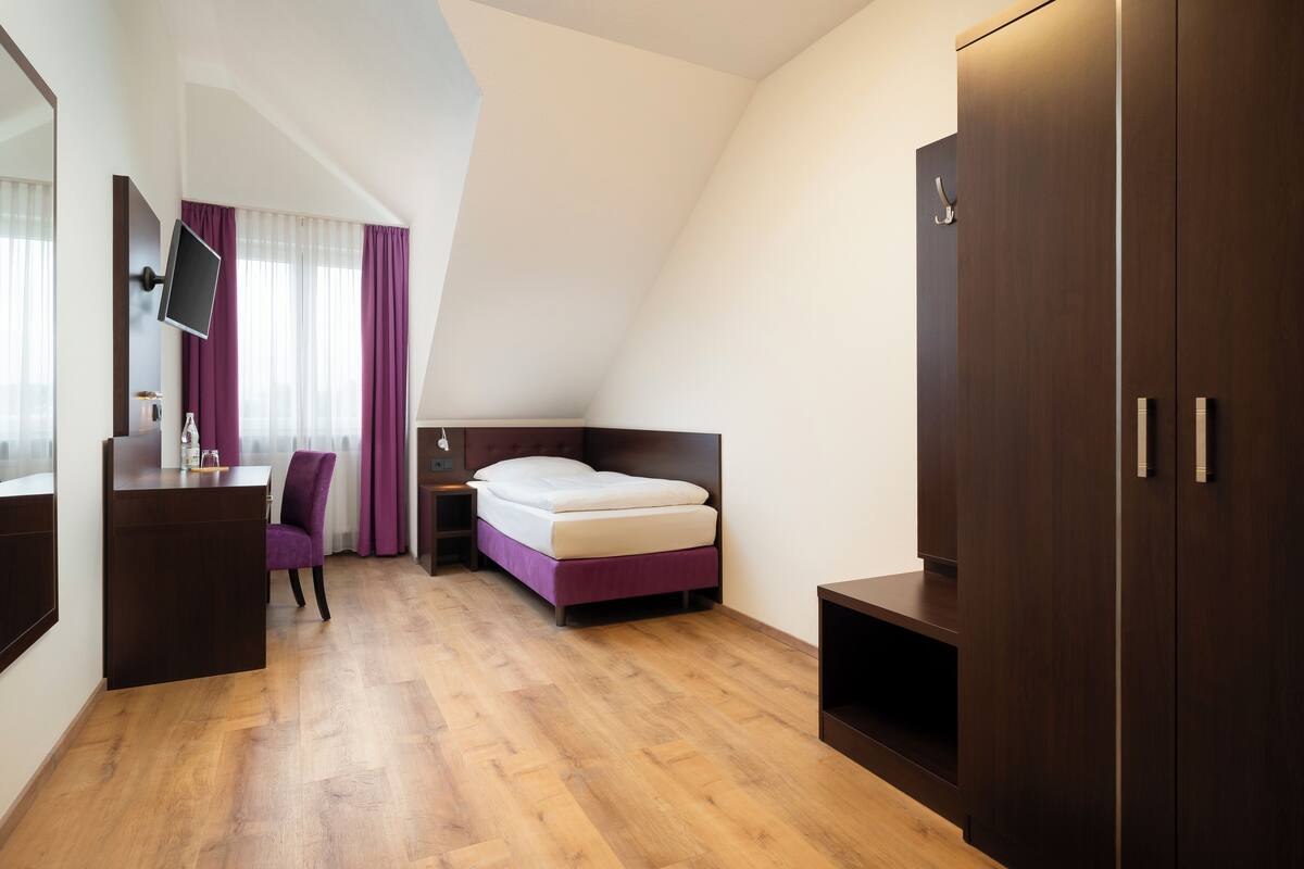 Quarto de hotel com cama, escrivaninha, cadeira, televisão e guarda-roupa.
