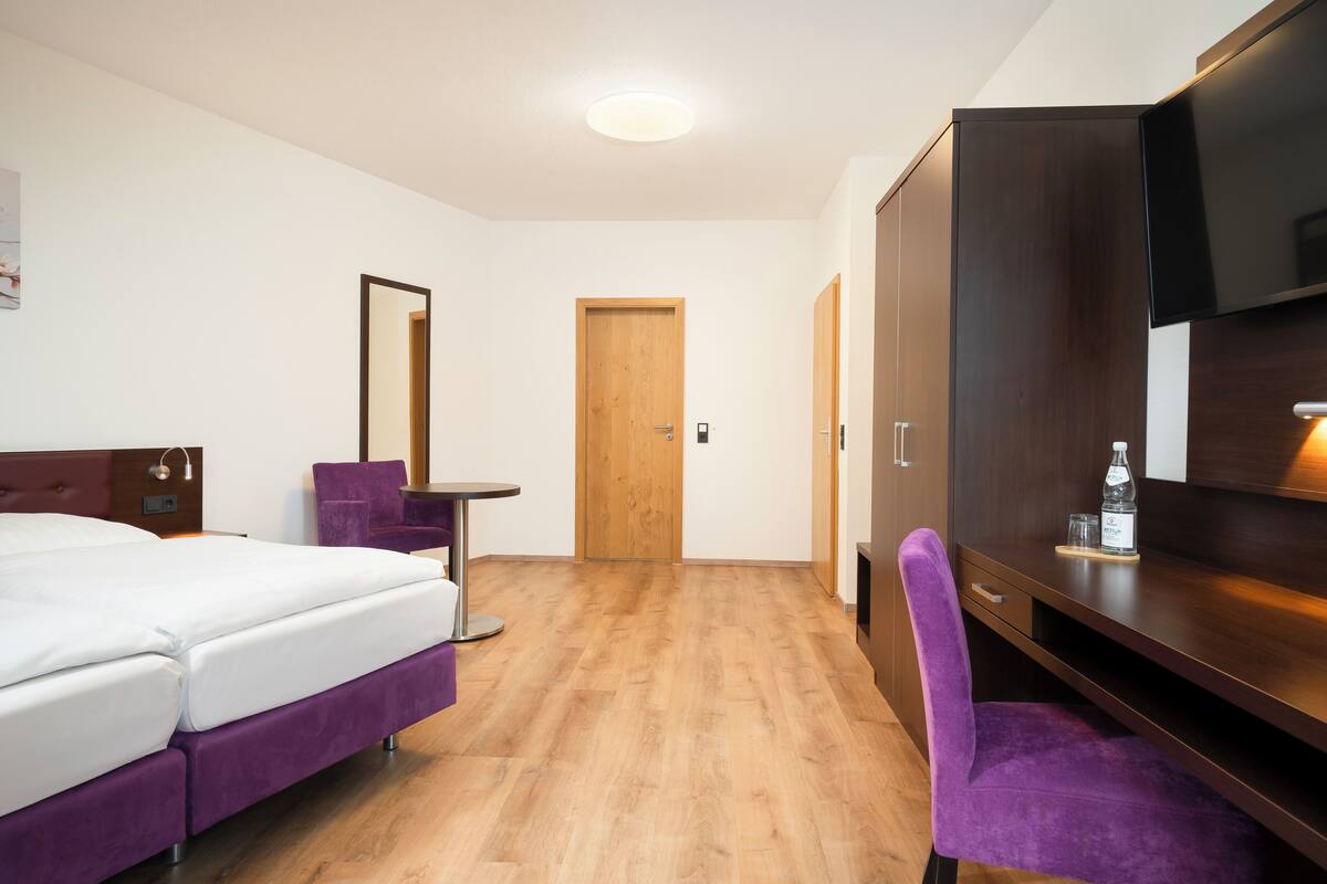 Quarto de hotel com cama, escrivaninha, cadeira, guarda-roupa e televisão.