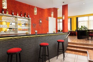 Zona de bar con taburetes, una barra con vasos y botellas, y una pared roja con relojes.