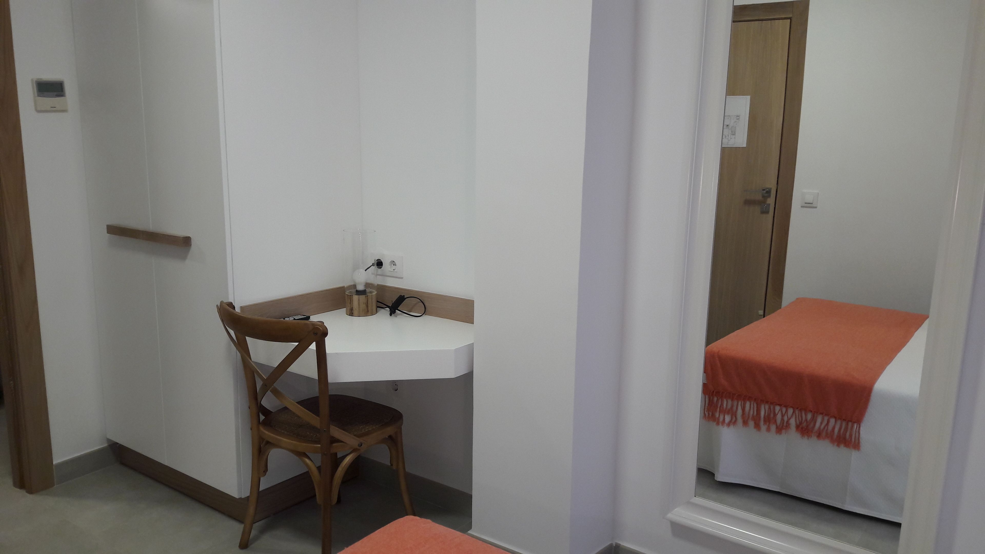 A small, clean room with a wooden chair, a white desk with a lamp, and a bed with an orange blanket.