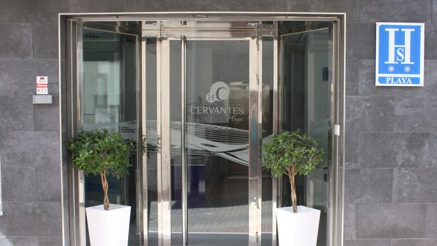 Entrada de un hotel con puerta de vidrio, dos plantas en macetas y un letrero que indica 'CERVANTES Playa'.
