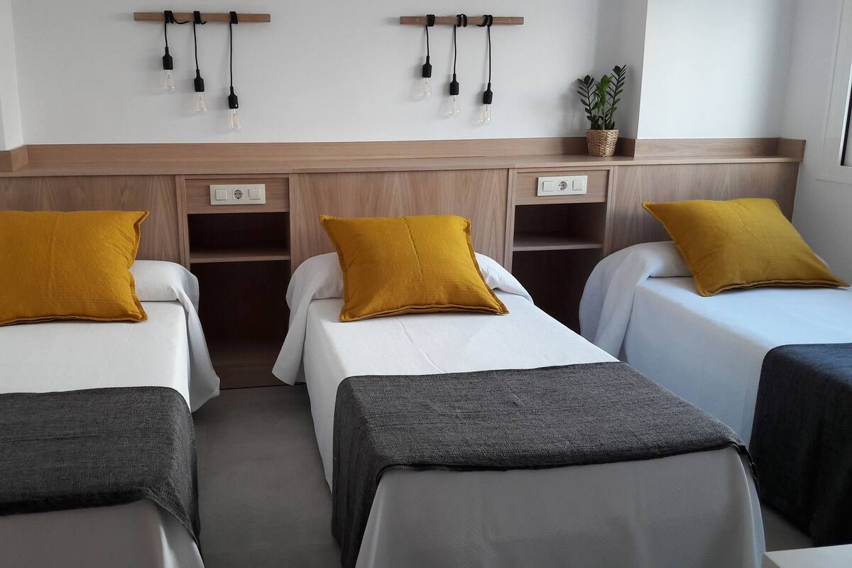 Habitación de hotel con tres camas individuales, cada una con sábana blanca y almohada color mostaza. El cabecero de madera tiene un estante sobre las camas que sostiene una planta y un juego de bombillas.