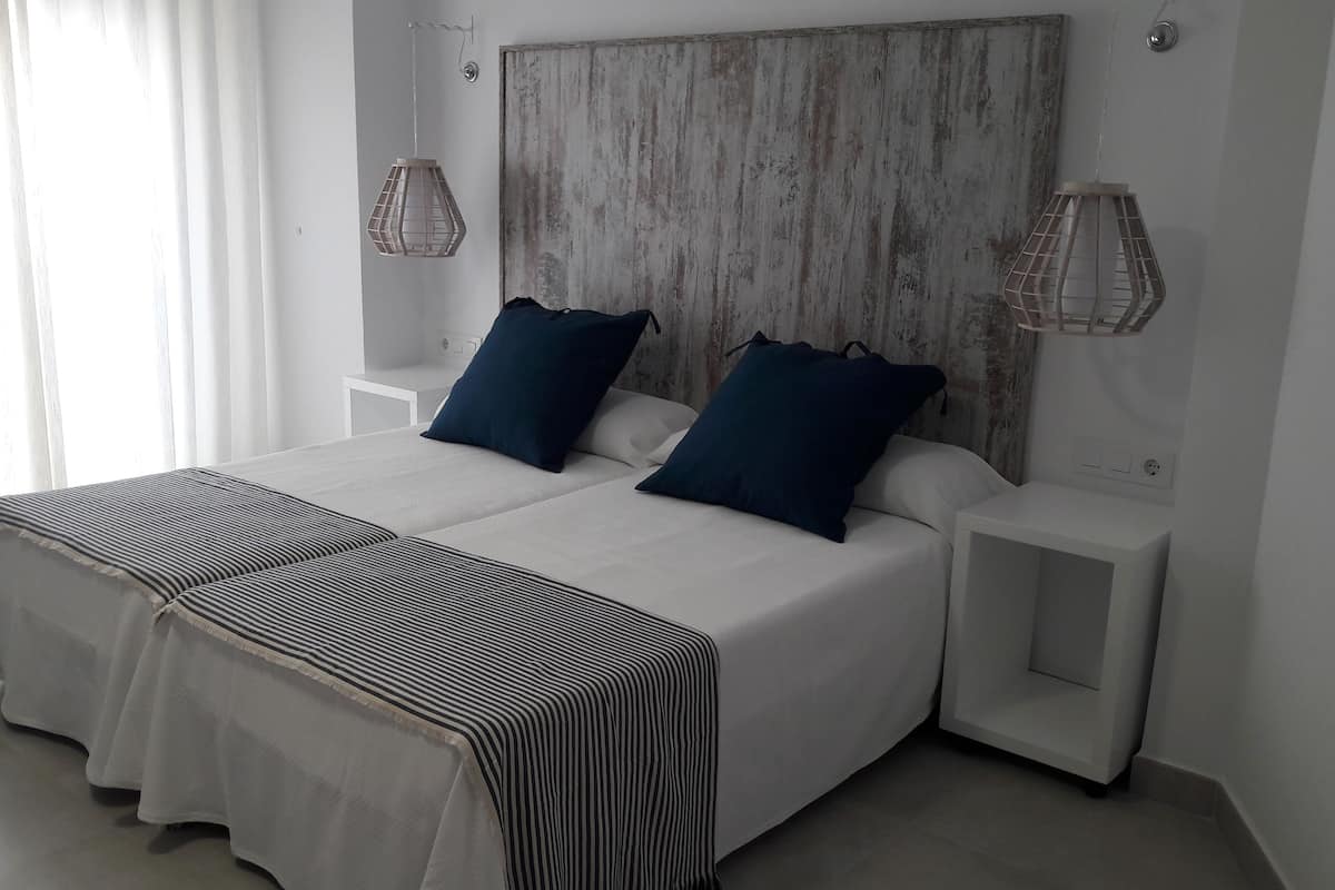 Una cama doble con cubrecama a rayas, dos almohadas oscuras y un cabecero de madera. Hay dos mesitas de noche con lámparas, y un ventanal con cortinas de gasa.