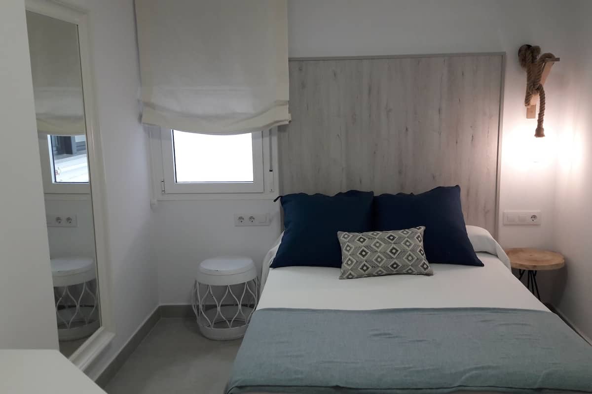 Un dormitorio con cama, una mesita de noche, un taburete pequeño y redondo, una ventana con persiana blanca y una lámpara fijada a la pared.