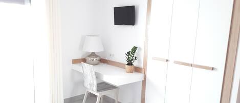Un escritorio blanco con una lámpara, una planta en maceta y un cubo de basura en una habitación con un armario blanco y un televisor montado en la pared.