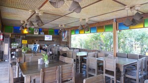 An indoor restaurant with wooden tables and chairs, hanging woven light fixtures, and a view of greenery outside. - Phuruarounmai Resort (Phu Ruea)
