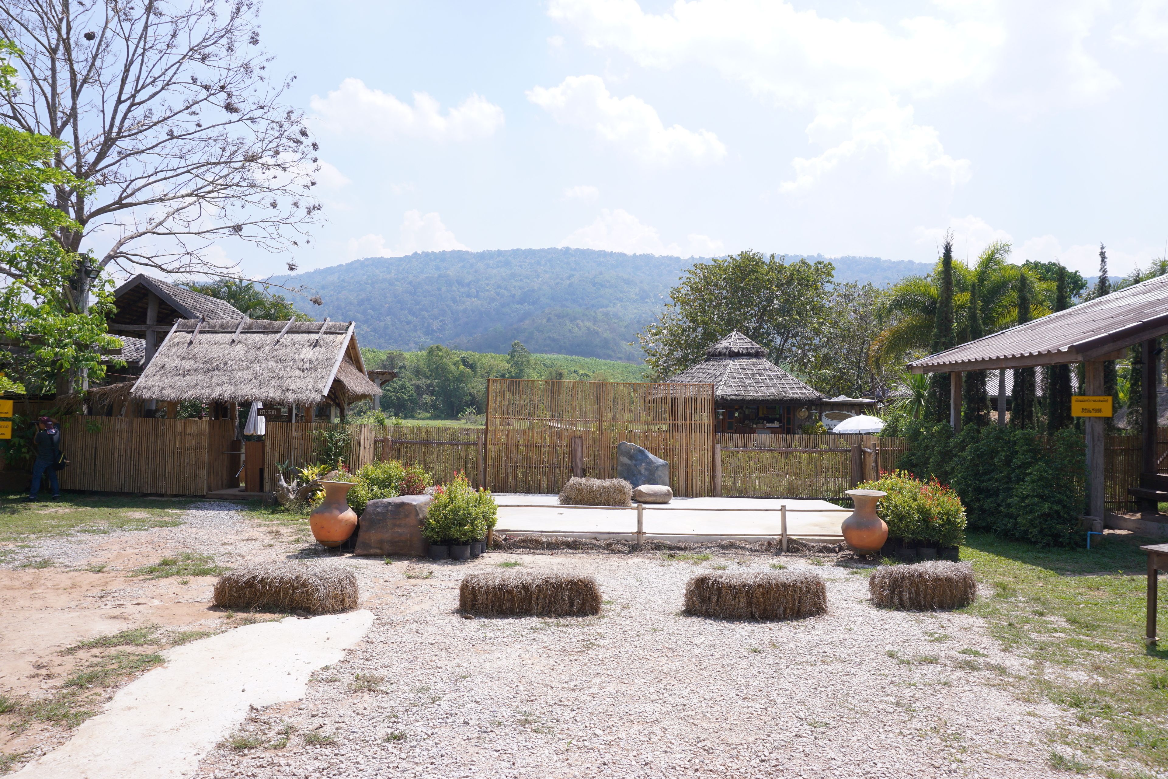 A rustic outdoor setting with thatched-roof buildings, hay bales, and a gravel path.